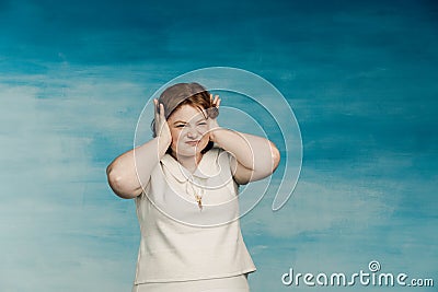 A plump young woman in a white dress with red hair covered her ears with her hands from annoying noise on a blue Stock Photo