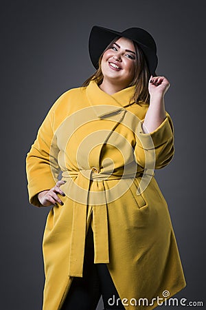 Plus size fashion model in yellow coat and black hat, fat woman on gray background, overweight female body Stock Photo