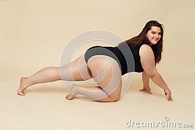 Plus Size Model. Fat Woman In Black Bodysuit Portrait. Crawling Brunette Posing On Beige Background. Stock Photo