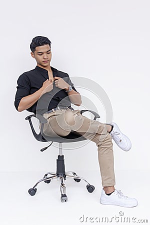 A poised young Filipino man dressed in smart casual clothes seated on an office chair, isolated on a white background Stock Photo