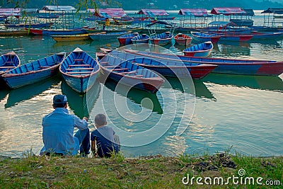 Pokhara, Nepal - September 04, 2017: Father and son sitting in the lakeshore enjoying the view of the boats in the lake Editorial Stock Photo