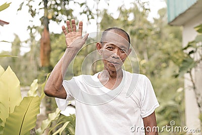 A poor but friendly old asian man waves his hand. A simple villager wearing a white shirt Stock Photo