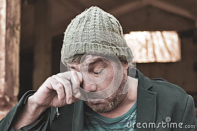 A poor homeless alcoholic with a swollen face in a hat and suit rubs his eyes with dirty hands crisis in modern society, the Stock Photo