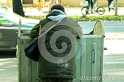 Poor and hungry homeless man in dirty clothes looking for food in the dumpster on the urban street in the city. Stock Photo