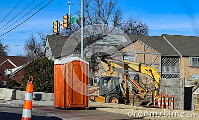 Portable bathroom at construction site at intersectio of urban roads with backhoe in background and traffic cones around Stock Photo