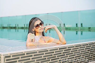 Portait of Beautiful Asian girl wearing sunglass with tropical cocktail and enjoying sunny weather at outdoor swimming pool. Stock Photo