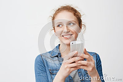Portait of cute tender redhead teenage girl with messy hair, looking aside and smiling while holding smartphone and Stock Photo