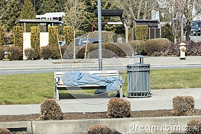 Portland, Oregon. Circa 2019. Homeless person covered with a blanket sleeping on a bench next to a garbage bin Editorial Stock Photo