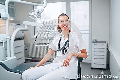 Portraait of sincerely smiling young dentist woman dressed white medical scrubs uniform sitting in modern dental clinic next to Stock Photo
