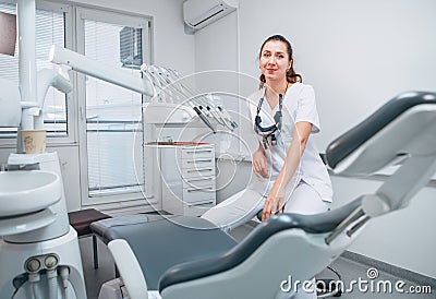 Portraait of sincerely smiling young dentist woman dressed white medical scrubs uniform sitting in modern dental clinic next to Stock Photo