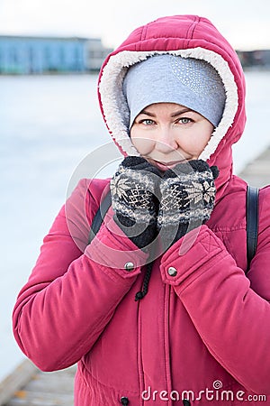 Portrait of an adult Caucasian woman dressed warm red jacket with hood on her head and knit mittens standing on quay of frozen Stock Photo