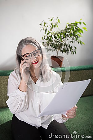 Portrait of an adult woman entrepreneur with grayish hair in a managerial position, dressed in a white blouse and busy working in Stock Photo