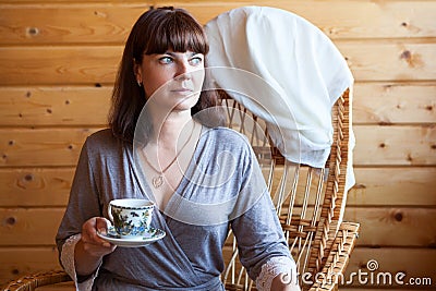 Portrait of an adult woman wearing gray bathrobe holding a cup of coffee, one person sitting rattan chair at home Stock Photo