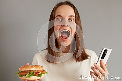 Portrait of amazed young adult caucasian woman wearing white t-shirt holding american classic fast food burger, using mobile phone Stock Photo