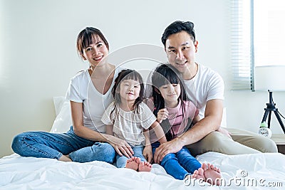 Portrait of Asian happy family smile and look at camera on bed at home. Loving couple and young little girl sibling daughters Stock Photo