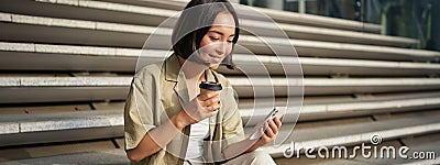 Portrait of asian woman with smartphone, drinks coffee and watches videos on mobile phone. Girl with telephone sits on Stock Photo