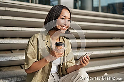 Portrait of asian woman with smartphone, drinks coffee and watches videos on mobile phone. Girl with telephone sits on Stock Photo