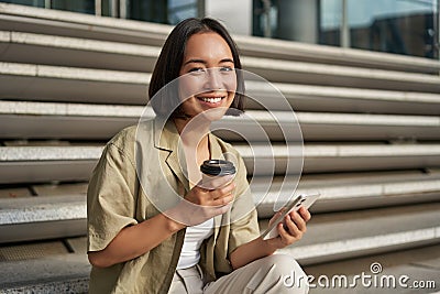 Portrait of asian woman with smartphone, drinks coffee and watches videos on mobile phone. Girl with telephone sits on Stock Photo