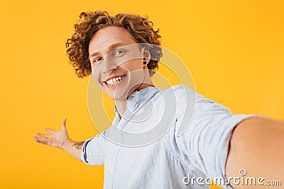 Portrait of astonished happy guy 20s taking selfie photo and holding copyspace on palm, isolated over yellow background Stock Photo