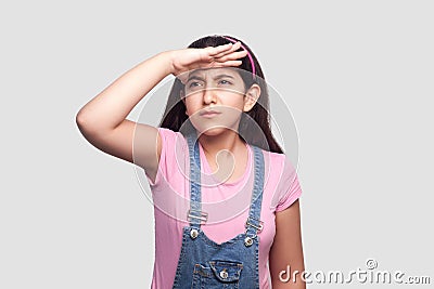 Portrait of attentive beautiful brunette young girl in casual pink t-shirt and blue denim overalls standing with hand on forehead Stock Photo