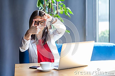 Portrait of attentive beautiful stylish brunette young woman in glasses sitting and looking at camera with crop composition hand Stock Photo