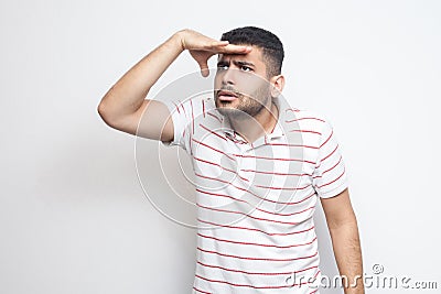 Portrait of attentive handsome bearded young man in striped t-shirt standing with hand on forehead and trying to look at something Stock Photo