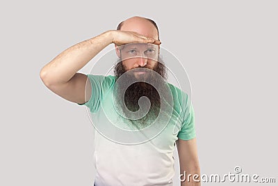 Portrait of attentive middle aged bald man with long beard in light green t-shirt standing with hand on forehead and looking far Stock Photo