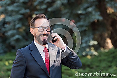 Portrait of attractive happy professional businessman dressed in suit and glasses talking on mobile phone in green park. Stock Photo