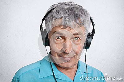 Portrait of attractive male pensioner has leisure time at home, listens music with modern headphones, looks directly at camera, lo Stock Photo