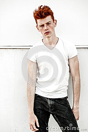 Portrait of attractive stylish young guy model with red hair and freckles standing near white wall , wearing white t-shirt. Fashio Stock Photo