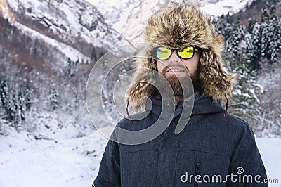 Portrait of an attractive young bearded man in a large fur hat and sunglasses against the snow-covered forest in the Stock Photo