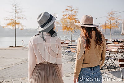 Portrait from back of brunette girl in hat talking with friend on nature background. Ginger lady in beige jacket Stock Photo