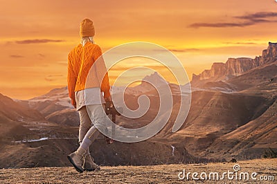 Portrait from the back of the girl traveler photographer in an orange sweater and hat with a camera in hand in the Stock Photo