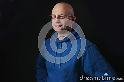 Portrait of bald and clean shaven man with glasses dressed in blue hoodie on a black background. Stock Photo