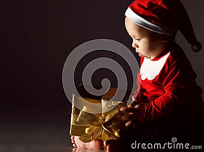 Portrait of barefooted infant baby boy in new year costume and red hat looking at his first new year gift golden box Stock Photo
