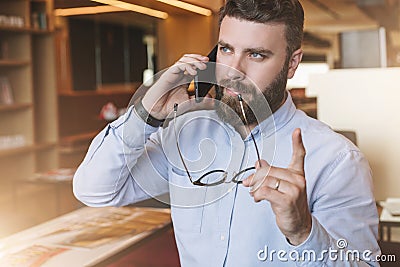 Portrait of bearded businessman in shirt,holding glasses in hand,lifting his index finger up, and talking on cell phone. Stock Photo