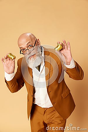 Portrait of bearded, mature, bald businessman in elegant, classical suit posing with golden coins against studio Stock Photo