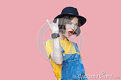 Portrait of beautiful amazed young woman in yellow t-shirt, blue denim overalls with makeup and black hat standing with rock horns Stock Photo