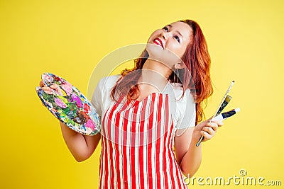 Portrait of a beautiful artist in an apron holding a palette and brush on a yellow background in the studio Stock Photo