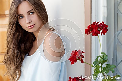 Portrait of a beautiful cute girl with blue eyes and dark curly hair in the courtyard near the wall with the window and flowers Stock Photo