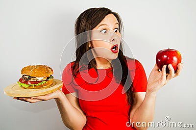 Portrait of a beautiful fat girl in surprise and holding a red apple and a hamburger, on gray background Stock Photo