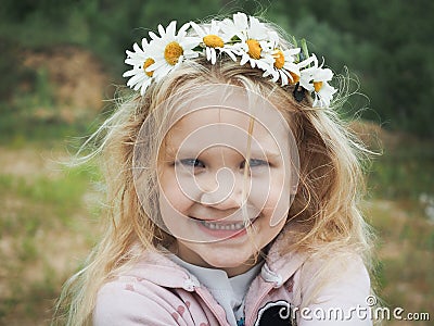 Portrait of a beautiful little girl with blond hair in a wreath of daisies. Field, green grass. Stock Photo