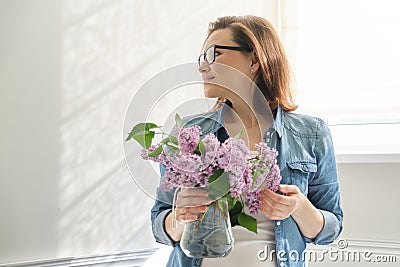 Portrait of beautiful mature woman at home with bouquet of lilac flowers. Background of home interior dining near the window Stock Photo