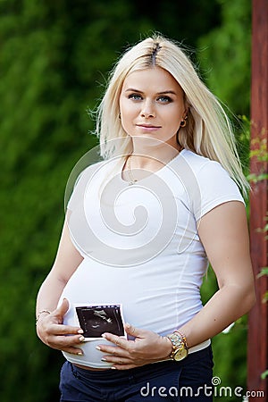 Portrait of beautiful pregnant woman in the flowering park. Young happy pregnant woman holds an ultrasonography photo Stock Photo