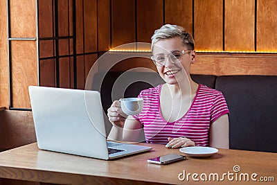 Portrait of beautiful satisfied young bussineswoman with blonde short hair in pink t-shirt is sitting in cafe and drinking coffee Stock Photo