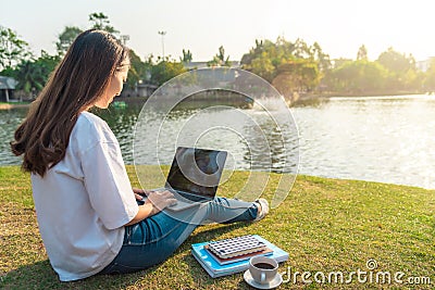 Portrait of beautiful smiling woman sitting on green grass in park with legs crossed during summer day and writing notes with pen Stock Photo