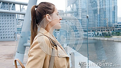 Portrait of beautiful smiling woman standing on bridge and looking on modern buildings and calm river Stock Photo