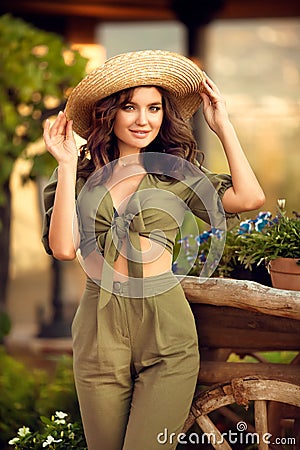 Portrait of beautiful woman smiling with curly hair in hat enjoying at park by wooden cart with green grass and flowers. Sunset Stock Photo