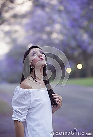 Portrait of beautiful woman in white dress standing in street surrounded by purple Jacaranda trees Stock Photo