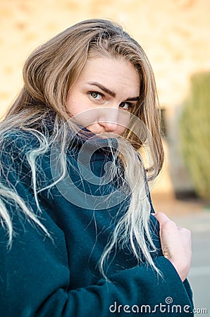 Portrait of beautiful young blonde girl in blue coat, who froze and hides in her collar, at sunset, against brick wall Stock Photo